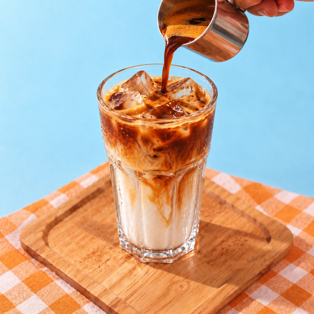 Espresso being poured from a small steel jug over an iced milk coffee in a tall glass