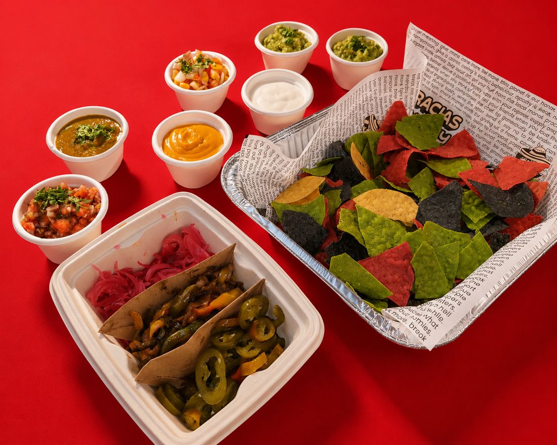 A foil tray of red, green, blue and yellow tortilla chips, seven cups of salsas, guacamole and dips, and a takeaway box of pickled onions, fajita vegetables and jalapenos on a red table