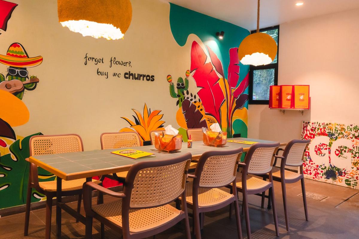Inside GG Chembur: cane chairs at a long table under yellow pendant lamps, beside a colourful mural that reads forget flowers, buy me churros